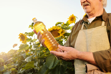 Senior farmer in apron and shirt looking at bottle of sunflower oil in his hands
