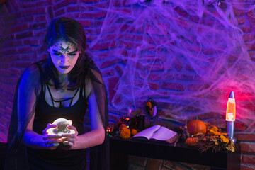 Halloween witch, young woman disguised as wicca holding a magic ball over spiderweb background with altar
