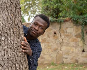 Primer plano de chico joven de raza negra, posando para una sesión fotográfica de retrato al aire libre, con camisa , abrazando un árbol y sonriendo. - obrazy, fototapety, plakaty