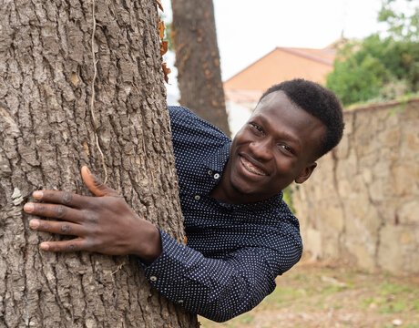 Primer plano de chico joven de raza negra, posando para una sesión fotográfica de retrato al aire libre, con camisa , abrazando un árbol y sonriendo.