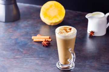 Pumpkin spice latte, coffee maker and milk jug on a dark background. Pumpkin latte with whipped cream and piece of pumpkin. Hot autumn drink