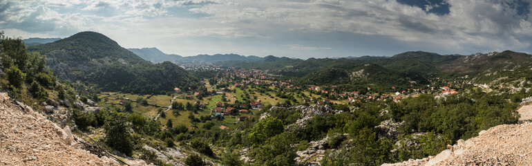 Fototapeta na wymiar Panorama of Cetinje, former capital city of Montenegro. View towards the houses of cetinje hiding in lush green hills and forests. Some rocks in the foreground.