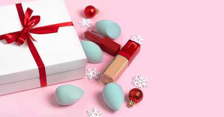 A set of decorative cosmetics makeup sponge, foundation, lipstick and snowflakes. White box with a red ribbon bow on a pink background with copy space. Concept for Valentine's Day, Christmas, New Year