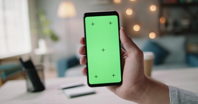 Hand of guys holding a smartphone with green chroma key screen in living room. Template for online store or social media 4k footage