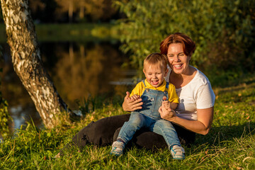 Fototapeta premium Mom and little boy sit on the river bank in the rays of the setting sun
