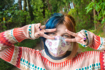 Funny portrait of a young woman with face mask, colorful hair and sweater, joking and playing with her hands (peace sign) while looking at camera. Green forest background and floral design mask.
