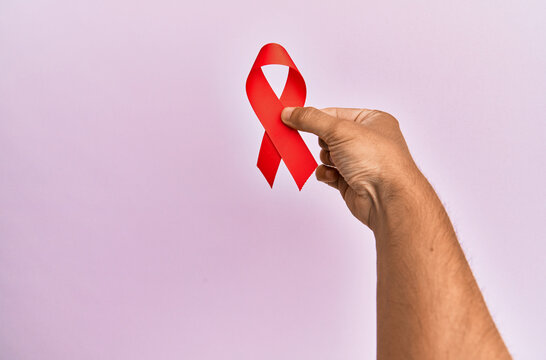 Hand of young hispanic man holding red ribbon over isolated pink background.