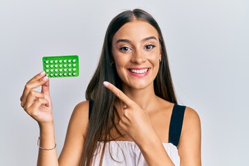 Young hispanic woman holding birth control pills smiling happy pointing with hand and finger