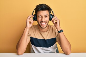 Young handsome man listening to music wearing headphones winking looking at the camera with sexy...