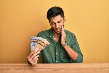 Young handsome man holding canadian dollars touching mouth with hand with painful expression because of toothache or dental illness on teeth. dentist concept.