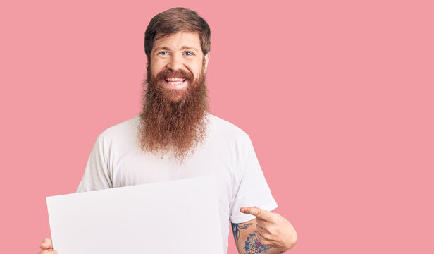 Handsome young red head man with long beard holding blank empty banner smiling happy pointing with hand and finger