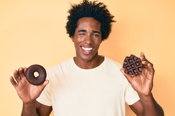 Handsome african american man with afro hair holding chocolate donut and waffle winking looking at...
