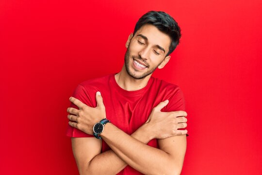Young handsome man wearing casual tshirt over red background hugging oneself happy and positive, smiling confident. self love and self care