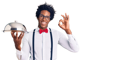 Handsome african american man with afro hair wearing waiter uniform holding silver tray doing ok sign with fingers, smiling friendly gesturing excellent symbol