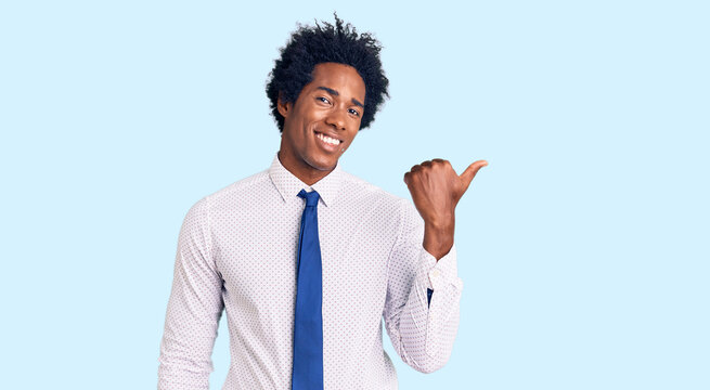 Handsome african american man with afro hair wearing business clothes smiling with happy face looking and pointing to the side with thumb up.