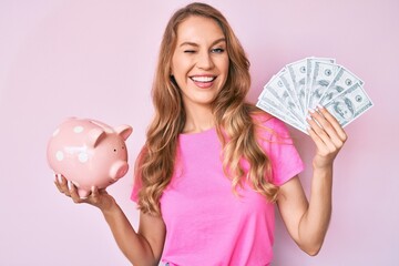 Young caucasian woman with blond hair holding dollars and piggy bank winking looking at the camera...