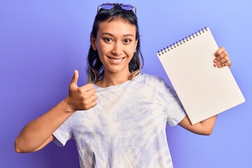 Young woman holding notebook smiling happy and positive, thumb up doing excellent and approval sign