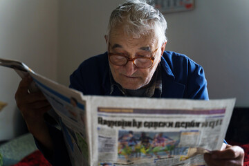 Front view of senior man reading newspapers at home in day - Old male pensioner peasant farmer sitting on bed at home reading press wearing eyeglasses - retirement leisure solitude real people concept