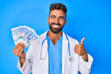 Young hispanic man wearing doctor uniform holding south african rand banknotes smiling happy and positive, thumb up doing excellent and approval sign