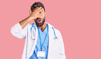 Young hispanic man wearing doctor uniform and stethoscope peeking in shock covering face and eyes with hand, looking through fingers with embarrassed expression.