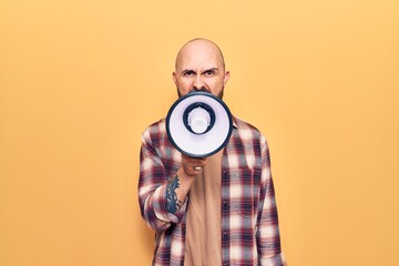 Young handsome bald man with angry expression. Screaming loud using megaphone standing over isolated yellow background