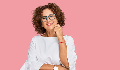 Beautiful middle age mature woman wearing casual clothes and glasses looking confident at the camera with smile with crossed arms and hand raised on chin. thinking positive.