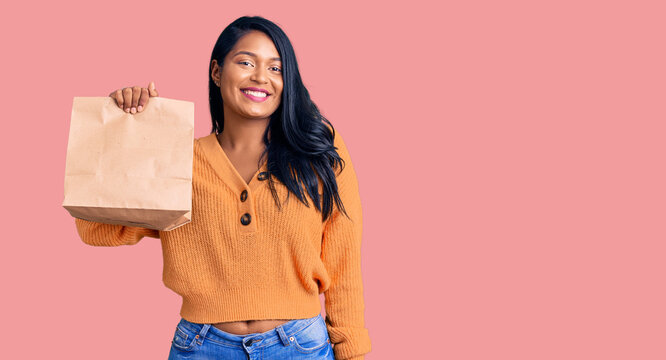 Hispanic woman with long hair holding take away paper bag looking positive and happy standing and smiling with a confident smile showing teeth