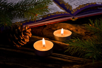 Two burning candles, an open book, fir branches and a pine cone on a wooden, old background. Concept for New Year and Christmas holidays, card, wallpaper.