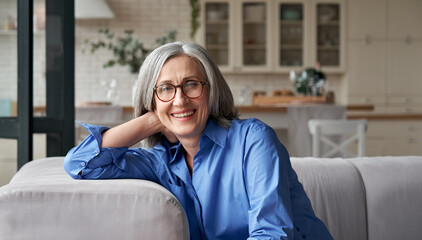 Happy relaxed mature old adult woman wearing glasses resting sitting on couch at home. Smiling...