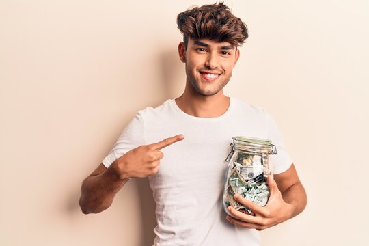 Young hispanic man holding jar with savings smiling happy pointing with hand and finger