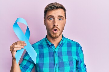 Handsome caucasian man holding blue ribbon scared and amazed with open mouth for surprise,...