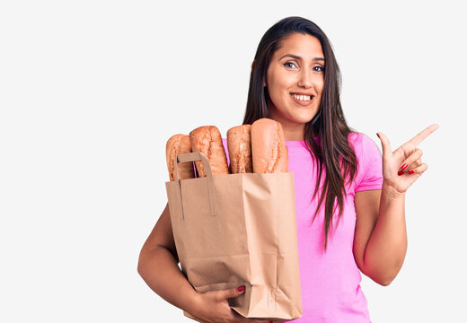 Young beautiful brunette woman holding delivery bag with bread smiling happy pointing with hand and finger to the side