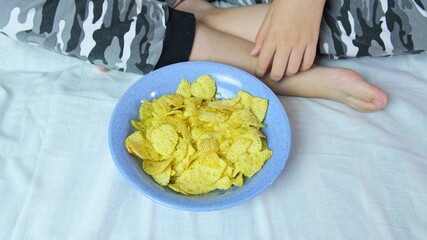 A boy sitting on a sofa at home and eating crispy potato chips from a bowl, close up view