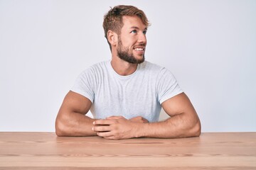 Young caucasian man wearing casual clothes sitting on the table looking away to side with smile on face, natural expression. laughing confident.