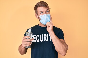 Young caucasian man wearing security t shirt and medical mask holding hand sanitizer gel serious face thinking about question with hand on chin, thoughtful about confusing idea
