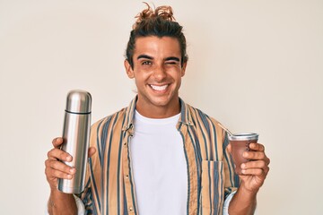 Young hispanic man drinking mate infusion winking looking at the camera with sexy expression,...