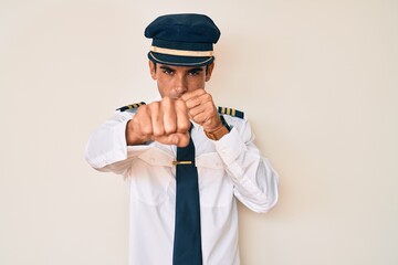 Young hispanic man wearing airplane pilot uniform punching fist to fight, aggressive and angry attack, threat and violence