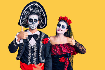 Young couple wearing mexican day of the dead costume over background doing happy thumbs up gesture with hand. approving expression looking at the camera showing success.