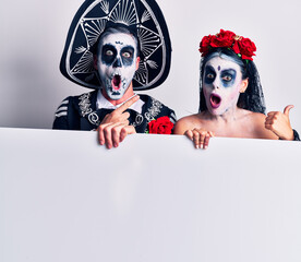 Young couple wearing mexican day of the dead costume holding blank empty banner surprised pointing with finger to the side, open mouth amazed expression.