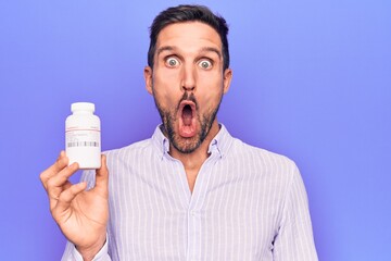 Young handsome man holding jar of medicine pills standing over isolated purple background scared and amazed with open mouth for surprise, disbelief face