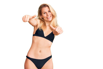 Young beautiful blonde woman wearing bikini approving doing positive gesture with hand, thumbs up smiling and happy for success. winner gesture.