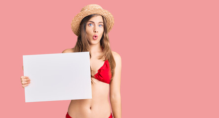 Beautiful caucasian young woman wearing bikini holding blank empty banner scared and amazed with open mouth for surprise, disbelief face