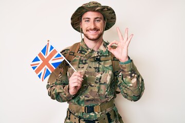 Young caucasian man wearing camouflage army uniform holding united kingdom flag doing ok sign with fingers, smiling friendly gesturing excellent symbol