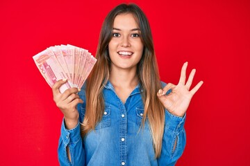 Beautiful caucasian woman holding yuan chinese banknotes doing ok sign with fingers, smiling friendly gesturing excellent symbol