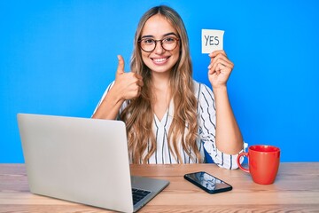 Young beautiful blonde woman holding yes reminder sitting on the table smiling happy and positive,...