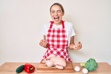 Fototapeta premium Young beautiful blonde woman wearing apron cooking chicken celebrating surprised and amazed for success with arms raised and eyes closed
