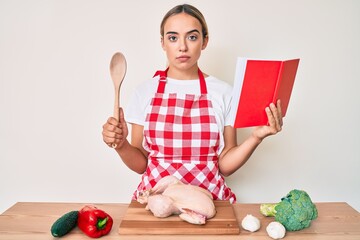 Young beautiful blonde woman wearing professional baker apron reading cooking recipe book relaxed with serious expression on face. simple and natural looking at the camera.