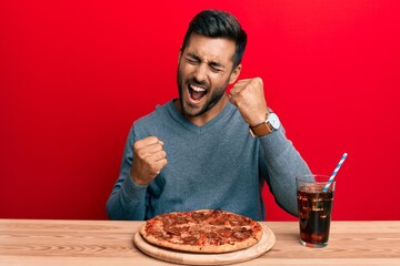 Handsome hispanic man eating tasty pepperoni pizza celebrating surprised and amazed for success...
