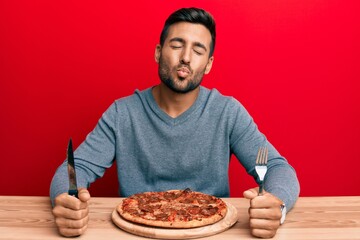 Handsome hispanic man eating tasty pepperoni pizza looking at the camera blowing a kiss being lovely and sexy. love expression.