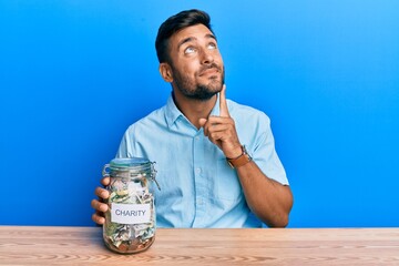 Handsome hispanic man holding charity jar with money serious face thinking about question with hand...